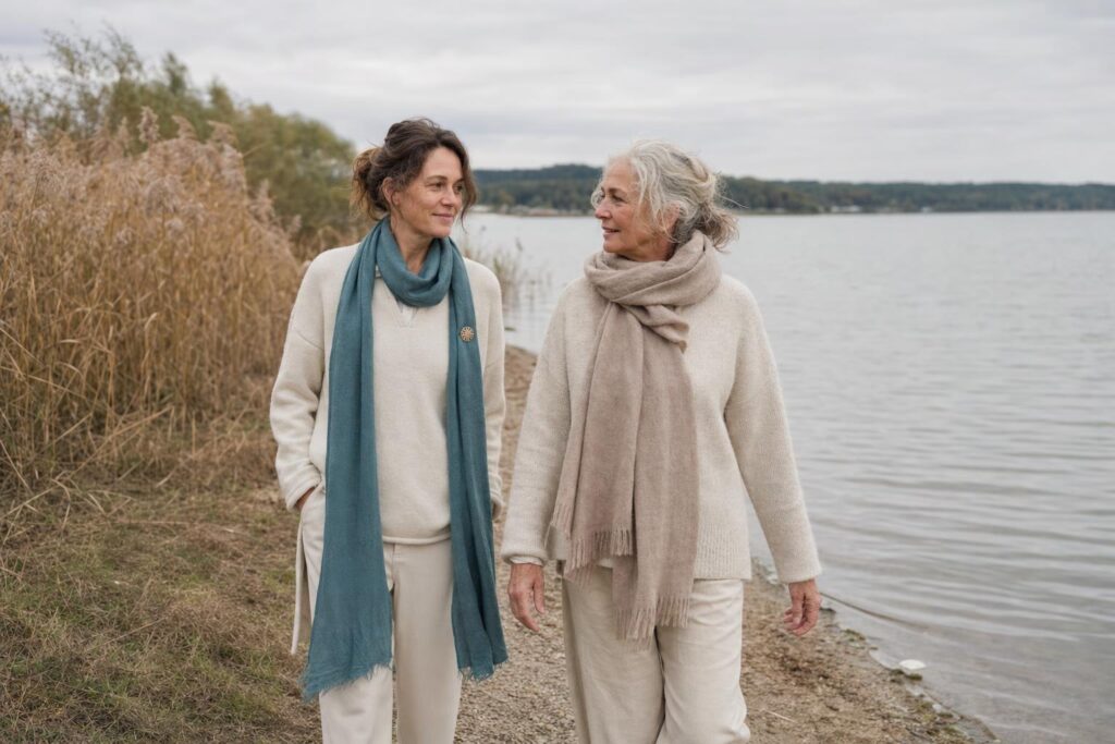 Zwei Frauen verschiedener Generationen gehen im Gespräch auf einem ruhigen Weg am Wasser.