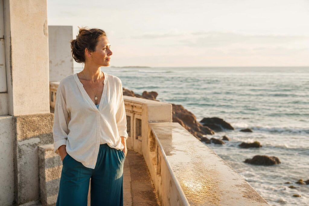 Frau in Creme und Petrol steht im goldenen Morgenlicht auf einer steinernen Terrasse am Meer.