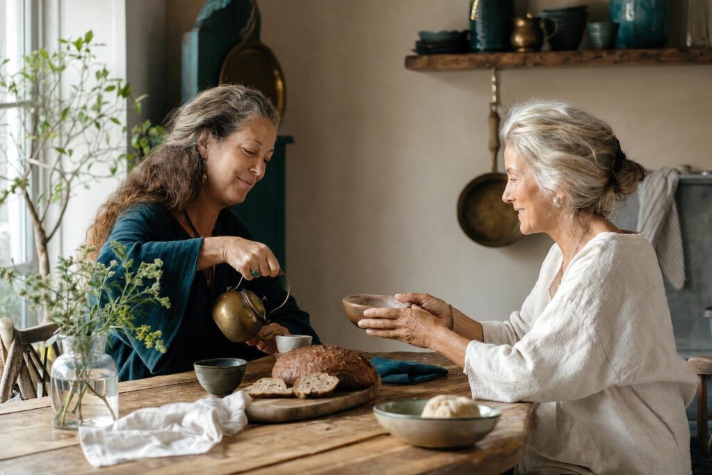 Zwei Frauen teilen in einer warm beleuchteten Küche Tee und eine Mahlzeit als Bild für gegenseitige Fürsorge.