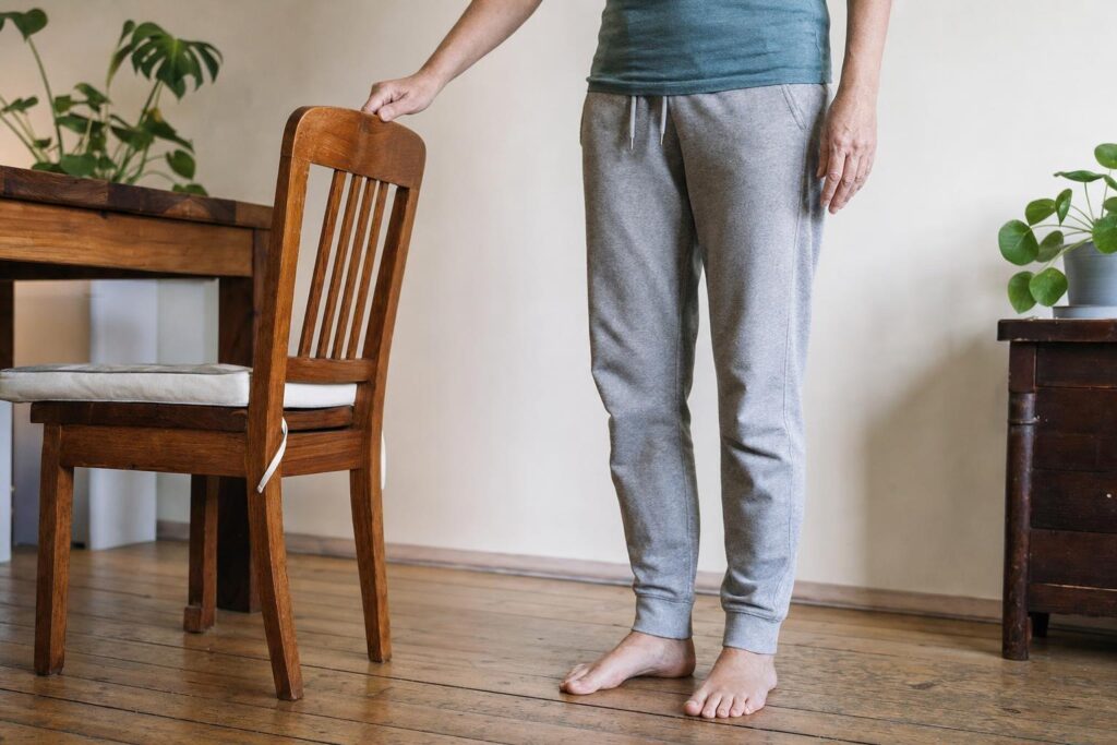 Person nutzt bei einer einfachen Standübung einen stabilen Stuhl als sichere Balancehilfe.
